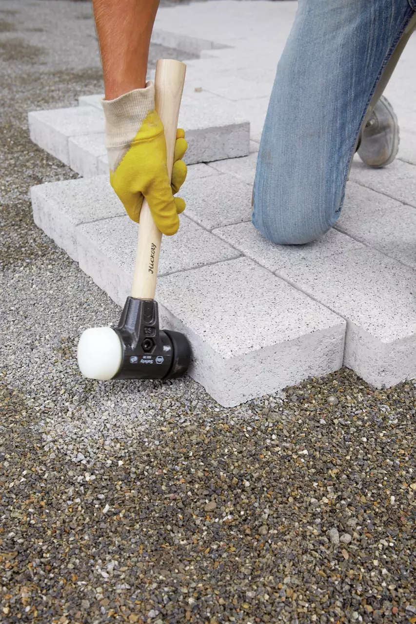 Person using a mallet with a wooden handle to lay paving stones on gravel.