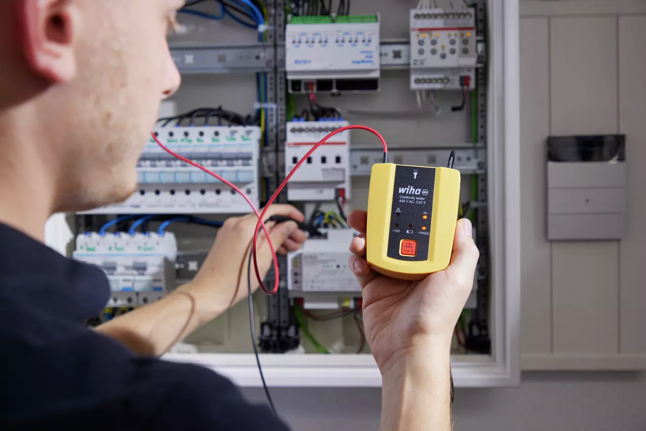 Person using a yellow continuity tester in front of an open electrical control cabinet.