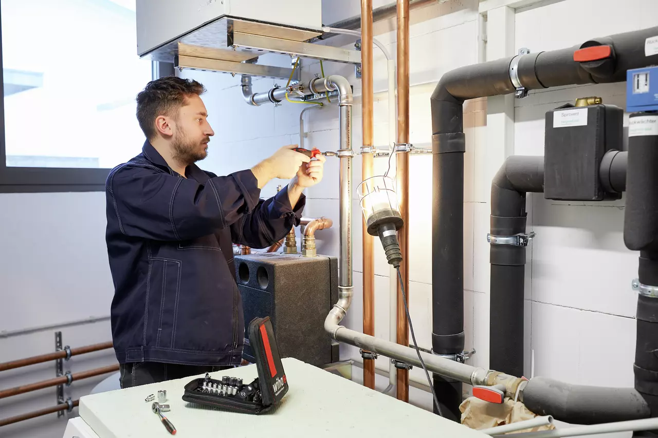 A person in a work jacket using a tool on pipes in a utility room. A tool set is visible in the foreground.