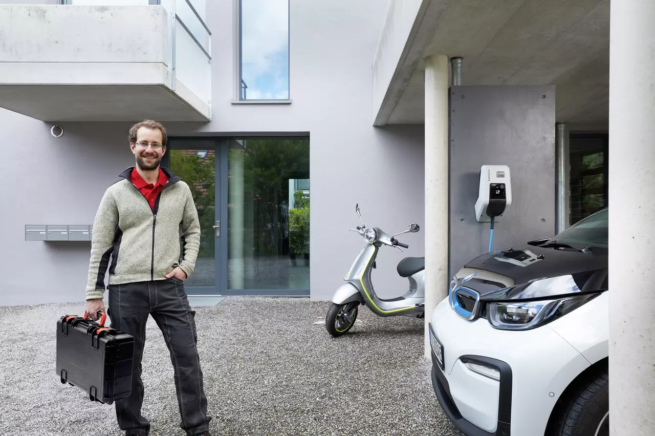 A person standing outside a modern building with an electric scooter and car charger nearby.