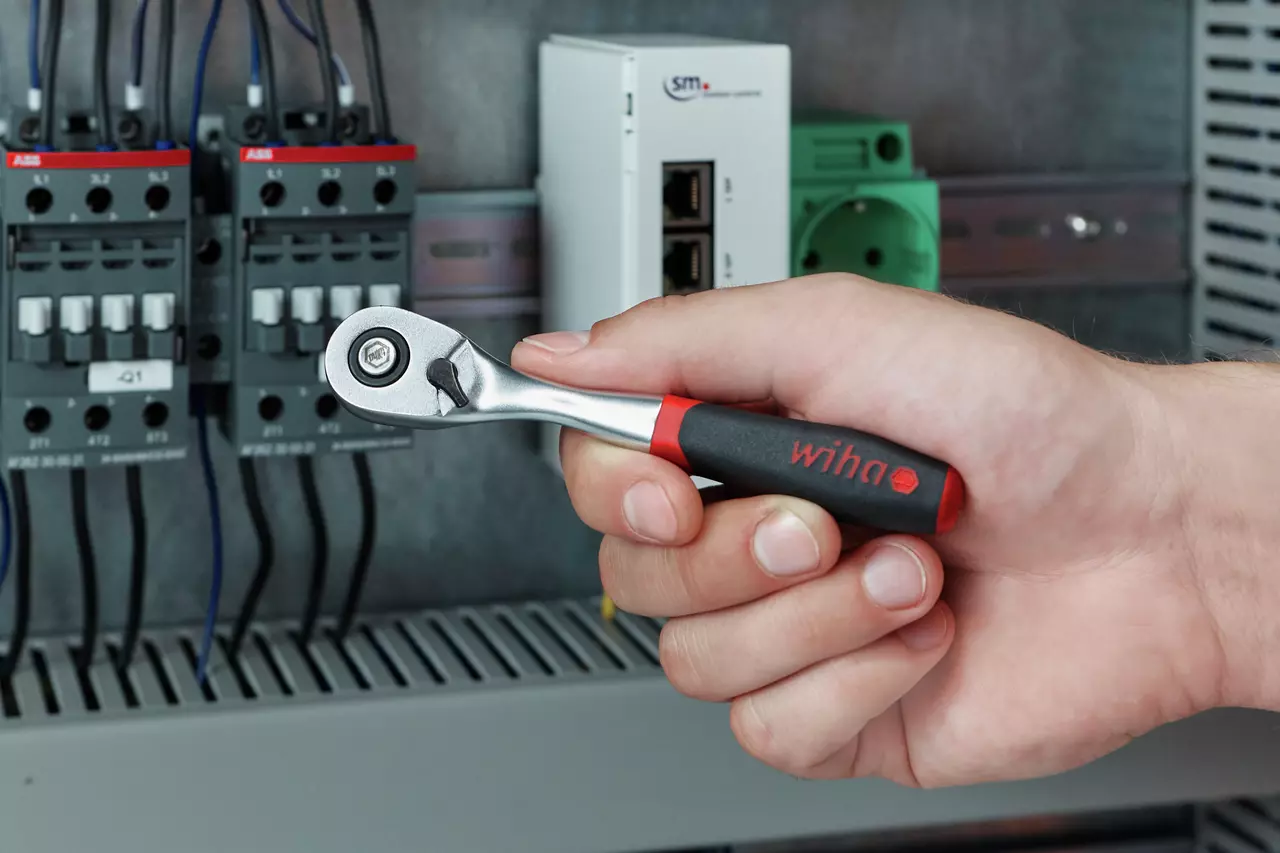 Hand holding a reversible ratchet with a black and red handle near electrical equipment.