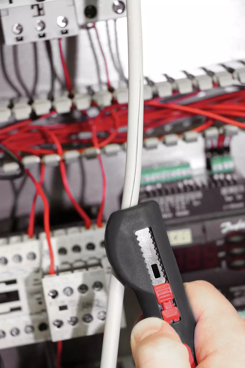 Hand using a multifunctional cable stripping tool on a white cable in front of an electrical panel.