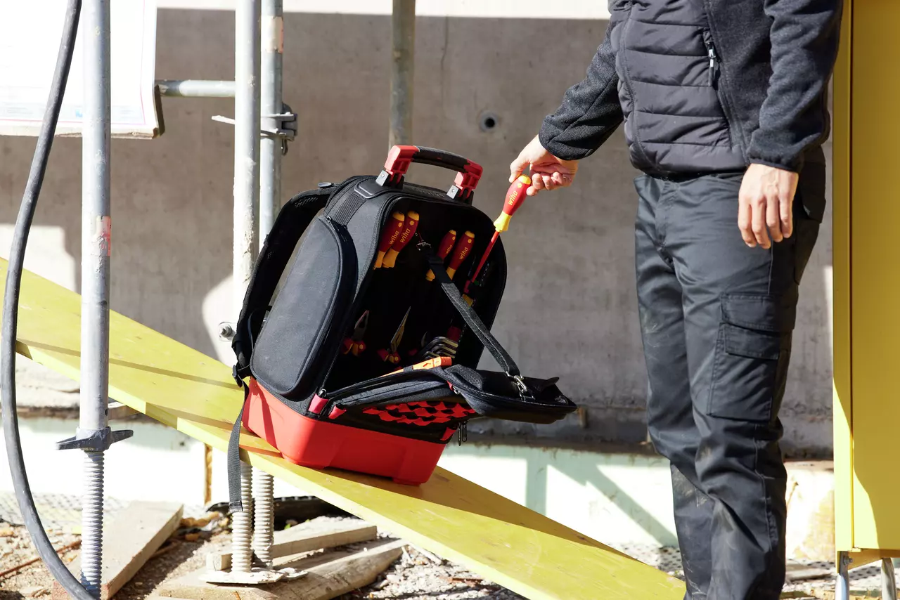 A person holding a screwdriver near an open tool bag on a construction site.