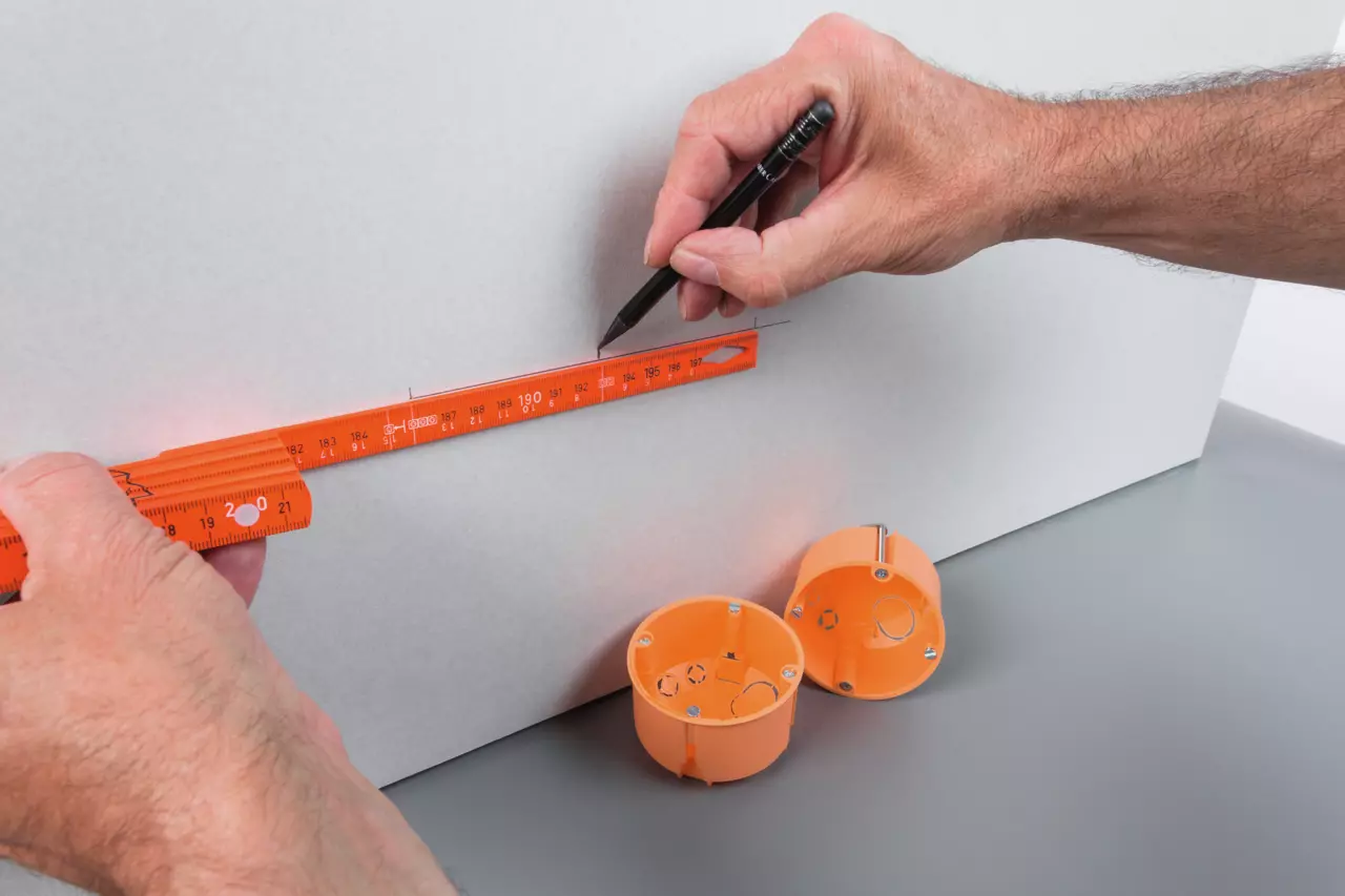 Person using an orange folding ruler to measure and mark on a white surface, with orange electrical boxes nearby.