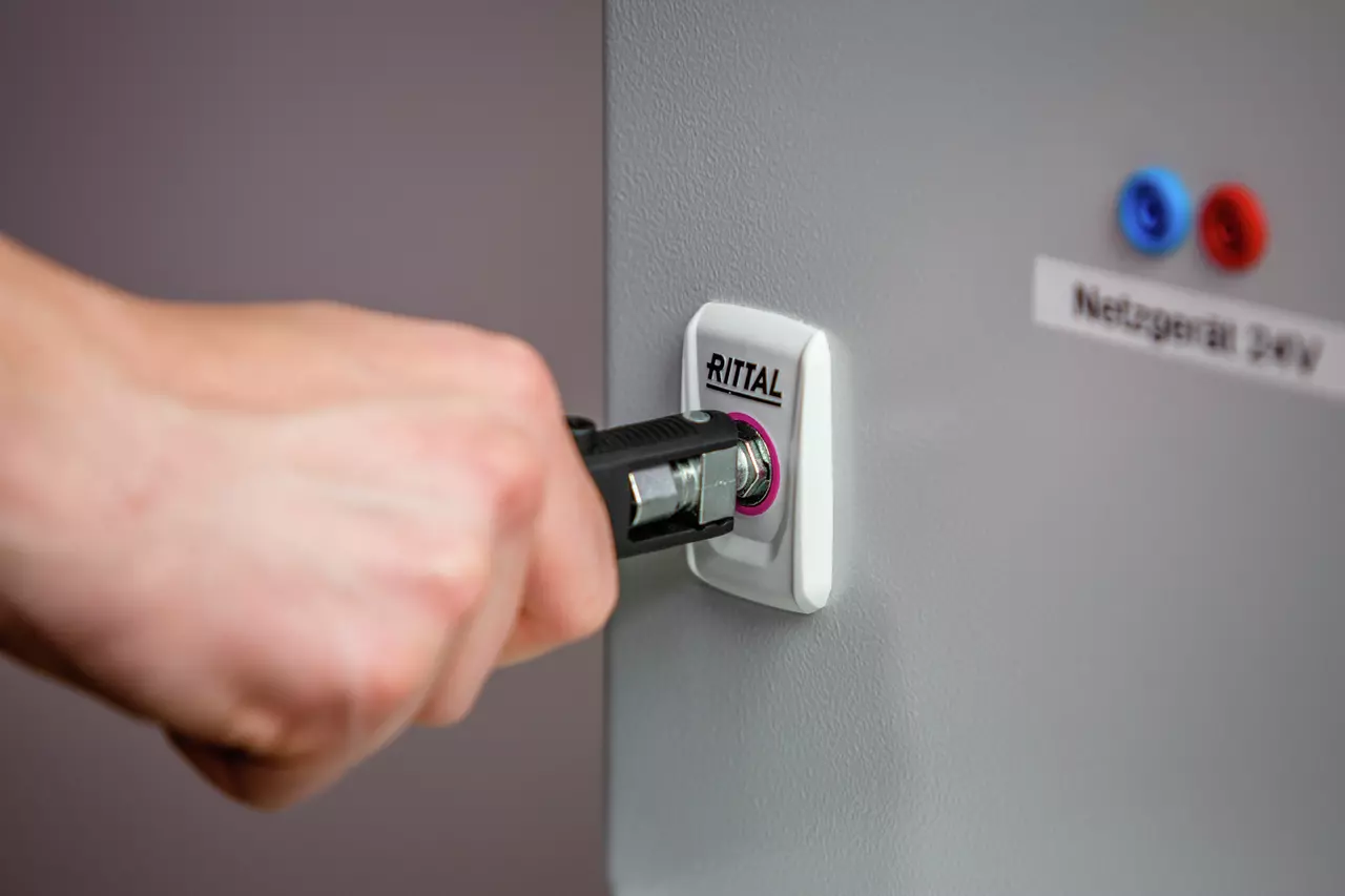 Hand using a tool to open a lock on a gray panel labeled "Rittal"