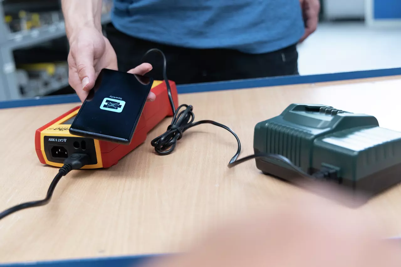 A person holds a smartphone next to a charging device on a table, with cables connected.