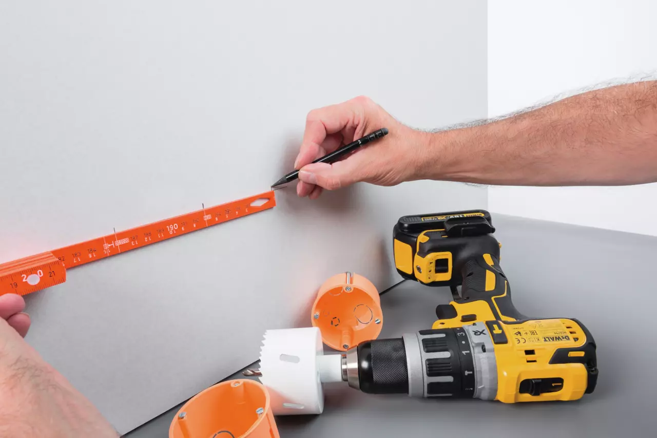 Person using a folding ruler and pencil on a wall, near a drill and orange boxes.