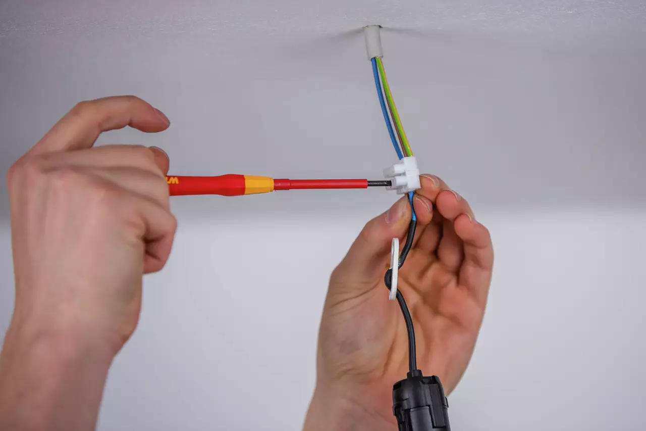Person using a red and yellow fine screwdriver on electrical wiring in the ceiling.