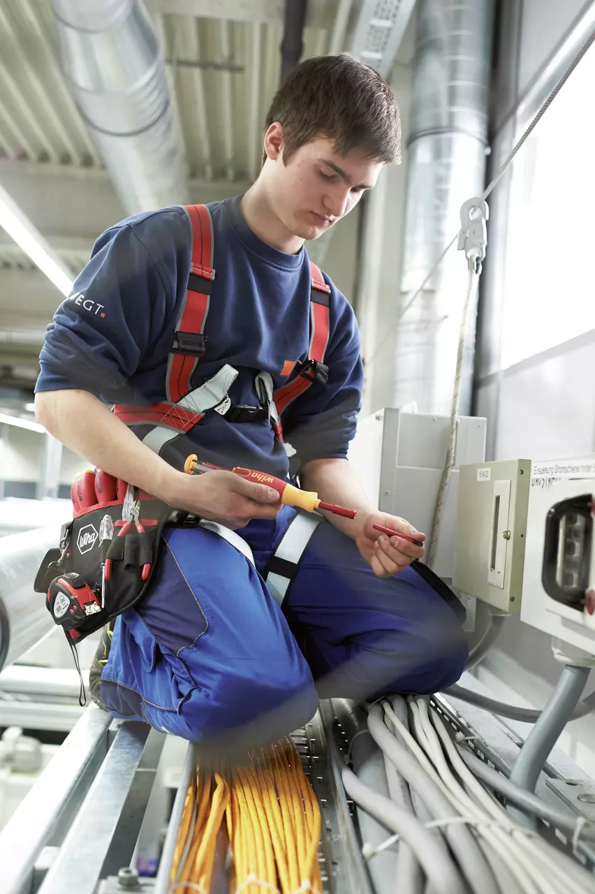 A person is using an electric screwdriver while kneeling on a metal structure with cables. They are wearing safety gear.