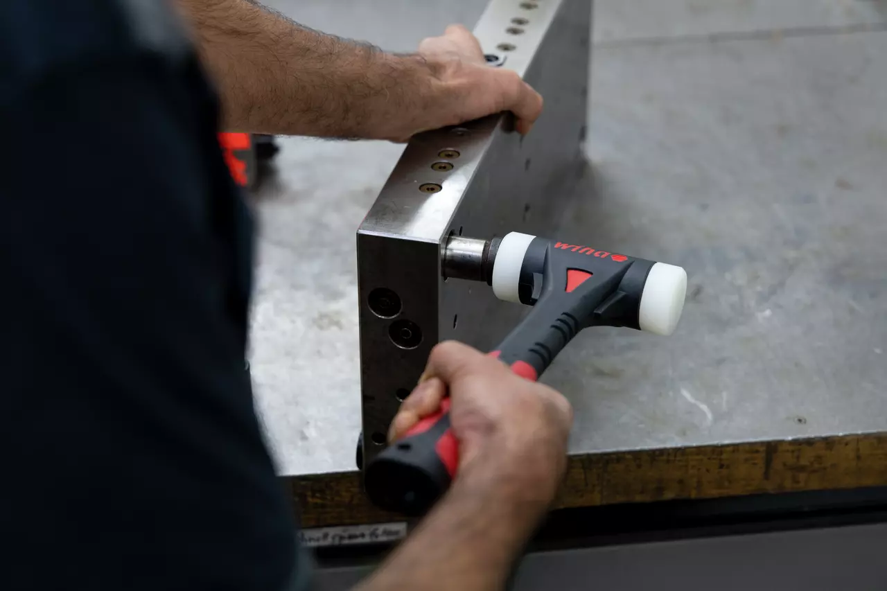 Eine Person benutzt einen Schonhammer an einem Metallblock auf einer Werkbank.