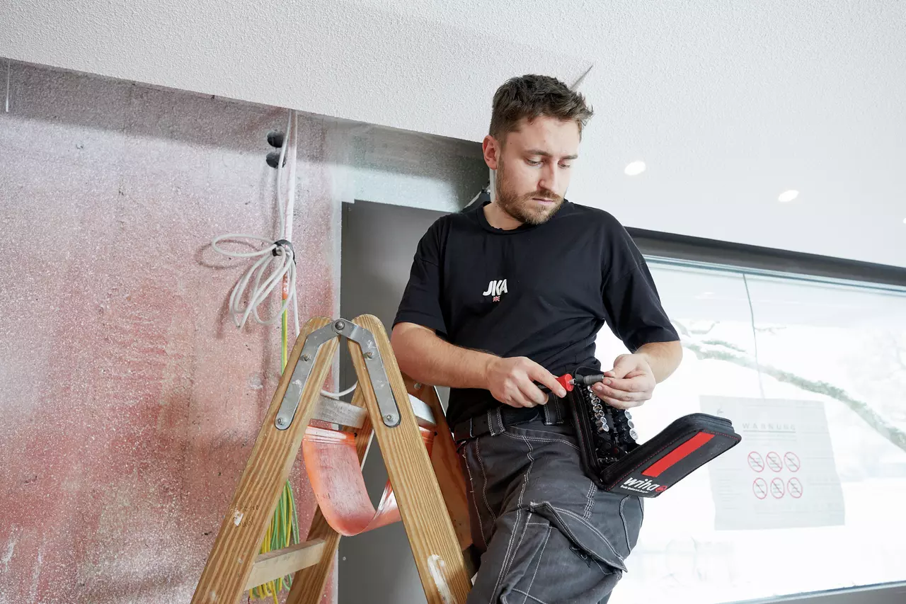 A person on a ladder holding a screwdriver set indoors.