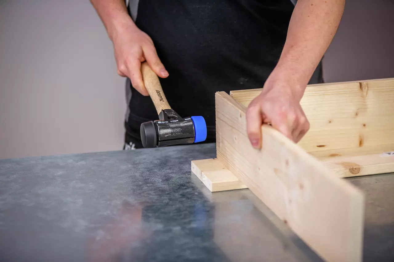 A person using a mallet with a blue rubber head to assemble wooden boards.