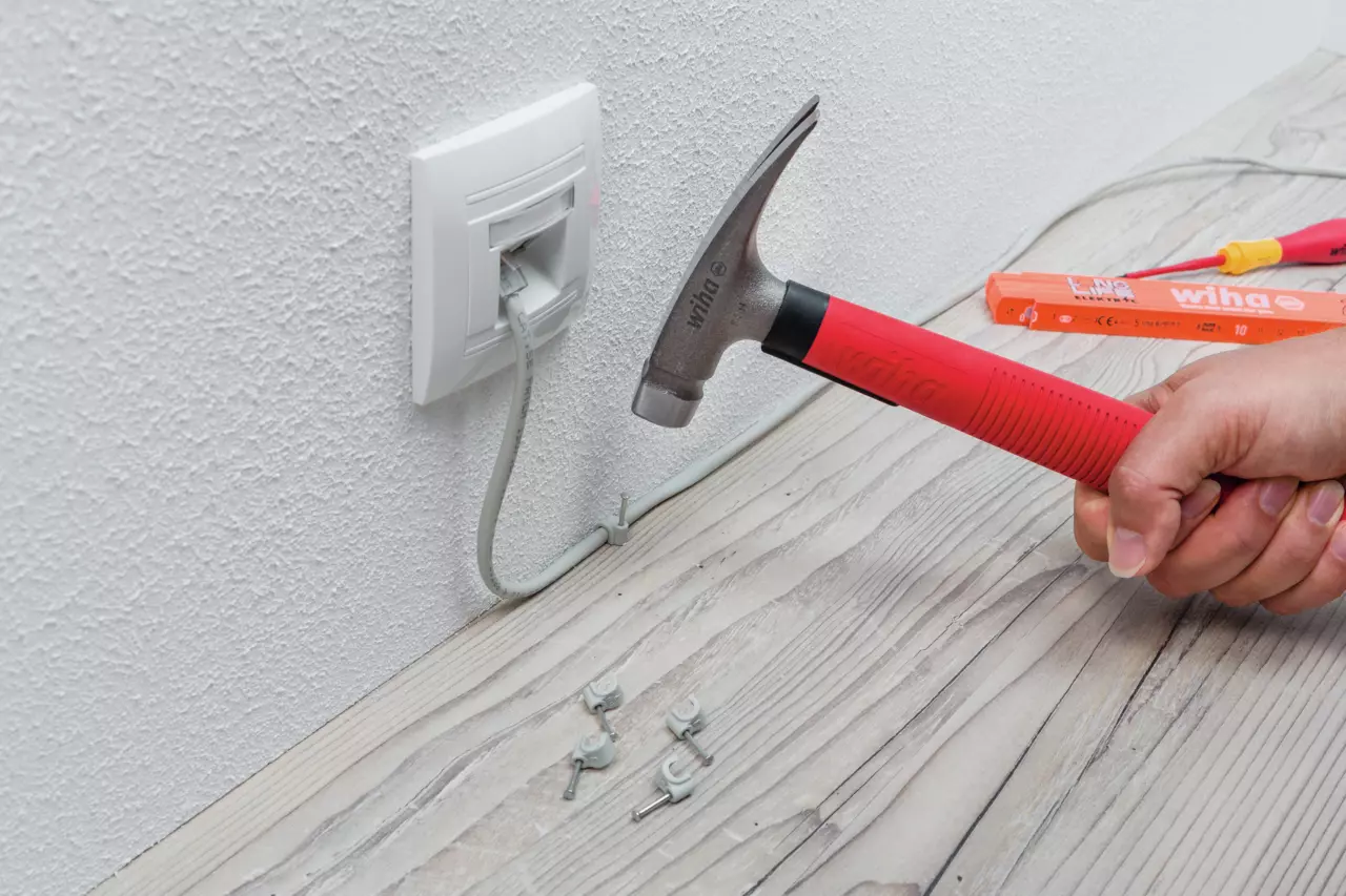 Electrician's hammer with red handle used to secure cable clips on a wooden surface near an outlet.