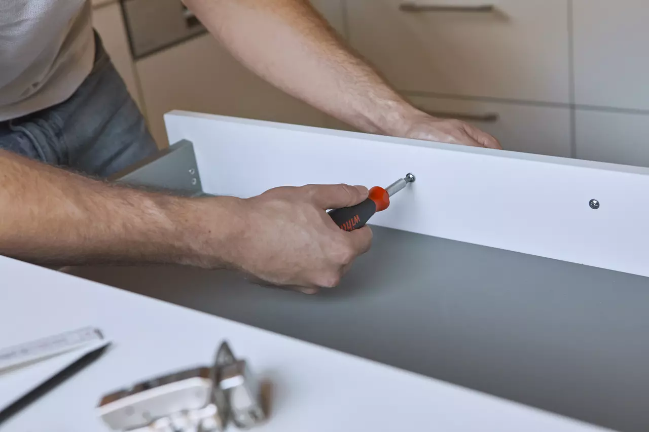Person using screwdriver on white drawer in kitchen