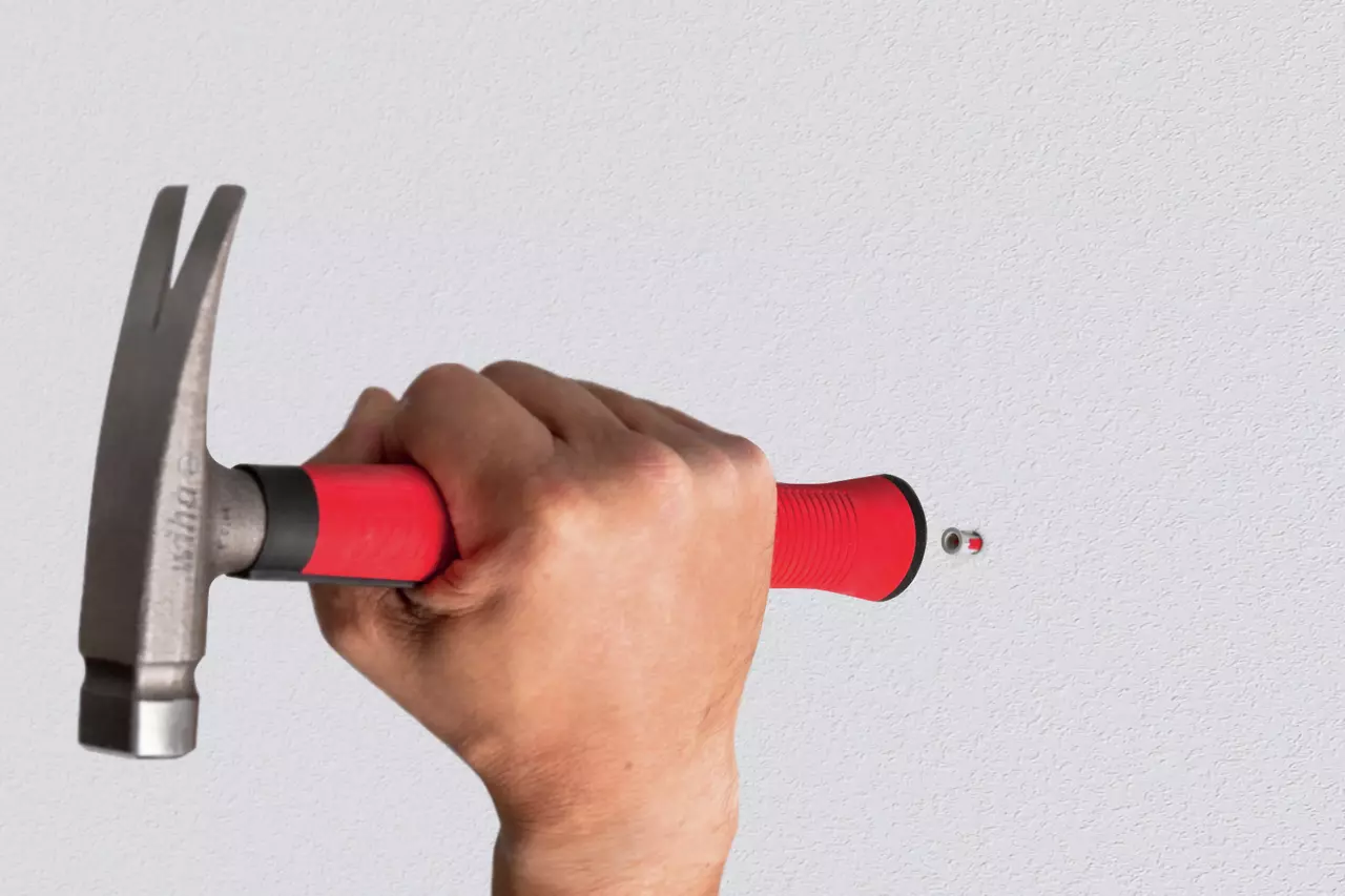 A hand holding a red-handled hammer against a gray wall.