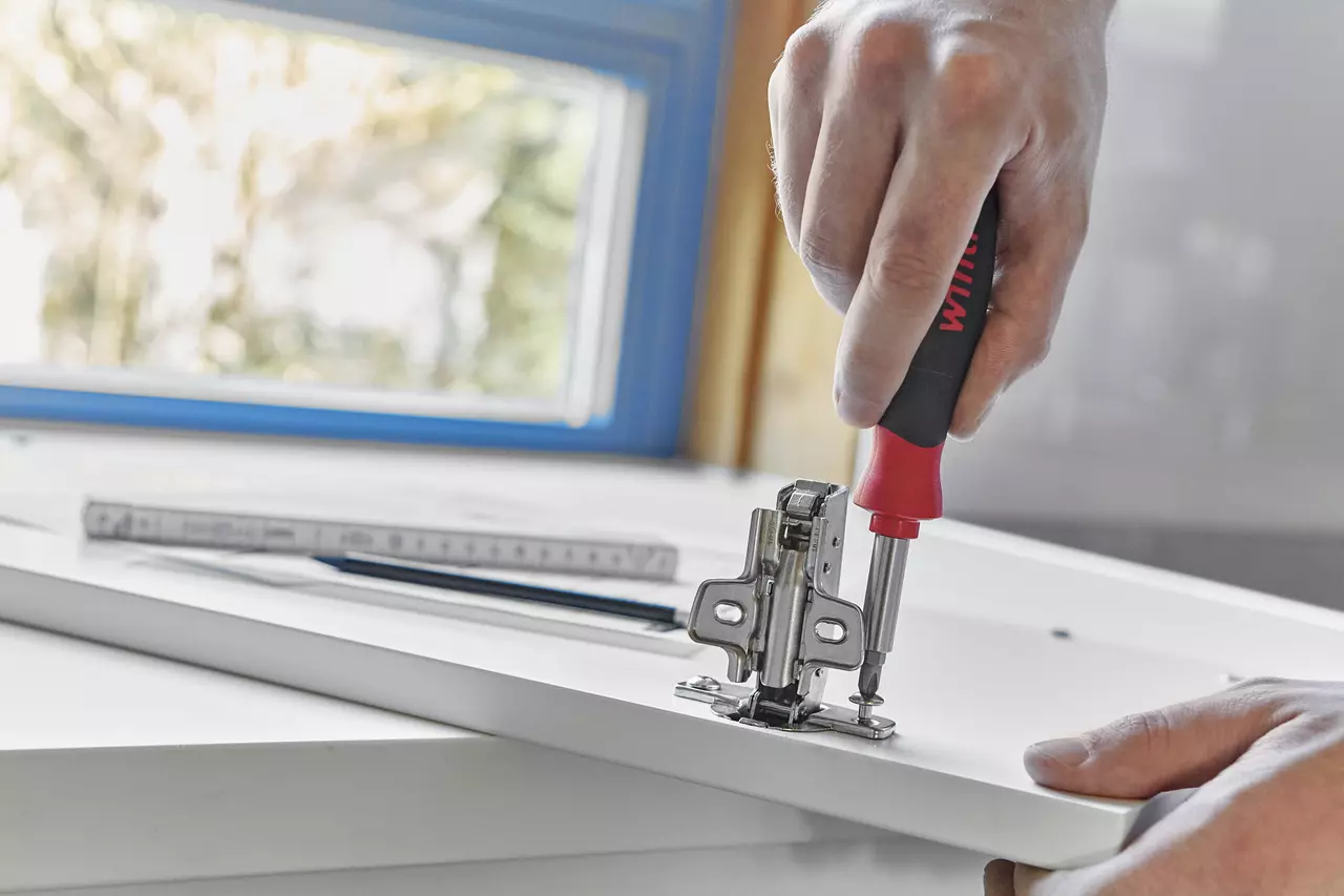 Person using a screwdriver to attach a hinge to a white cabinet door in a kitchen.