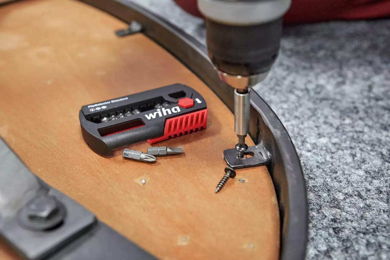 A drill driving a screw into a bracket on a wooden surface next to a bit set.