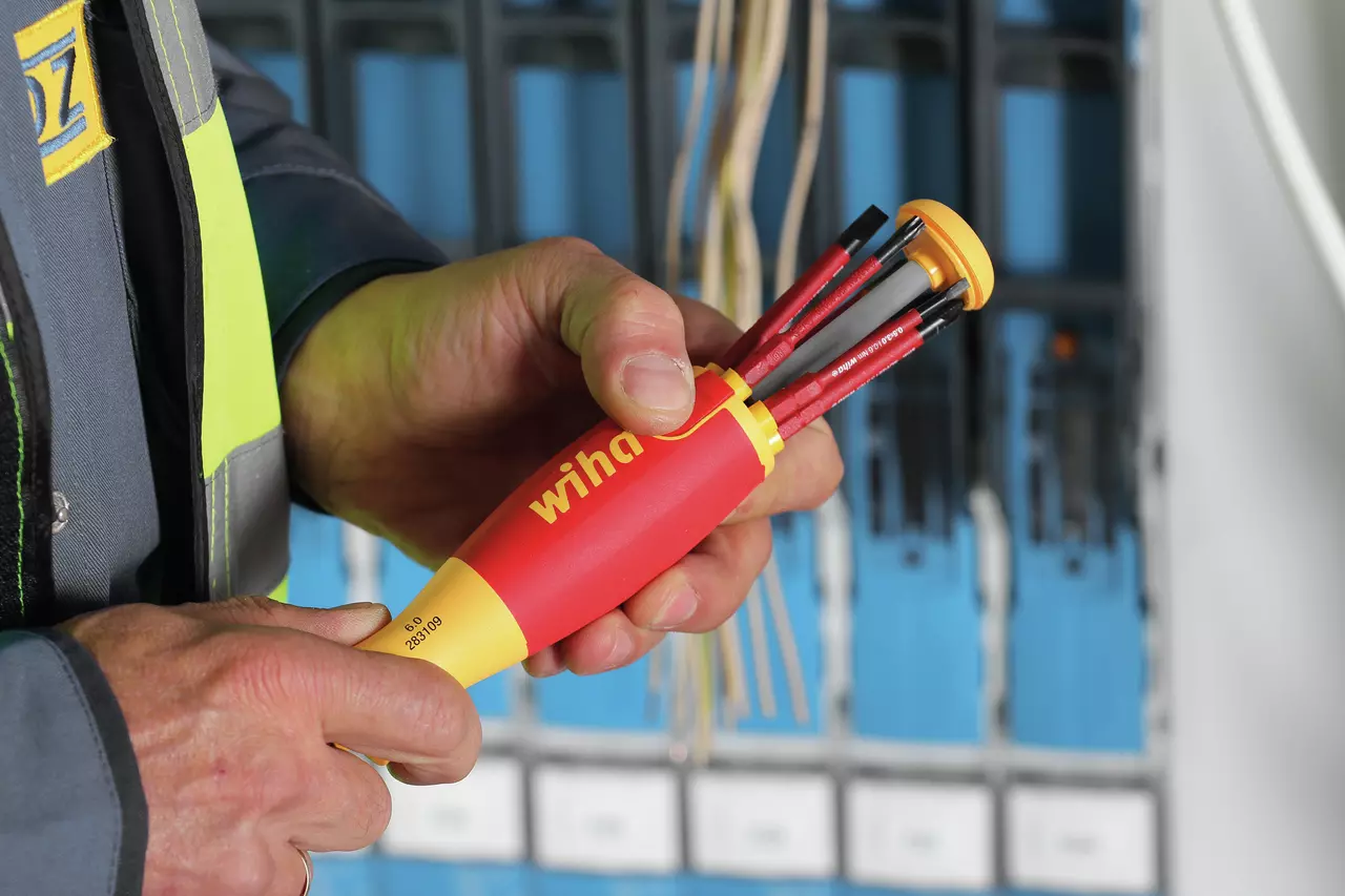 A person holding a red and yellow electric screwdriver with multiple bits in front of a circuit panel.