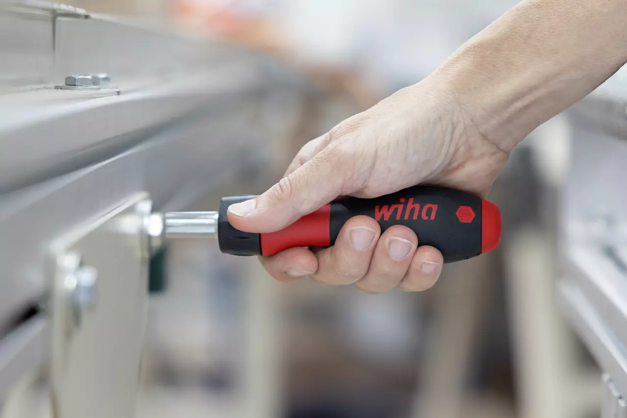 Hand holding a ratchet screwdriver with a black and red handle, working on a metal surface.