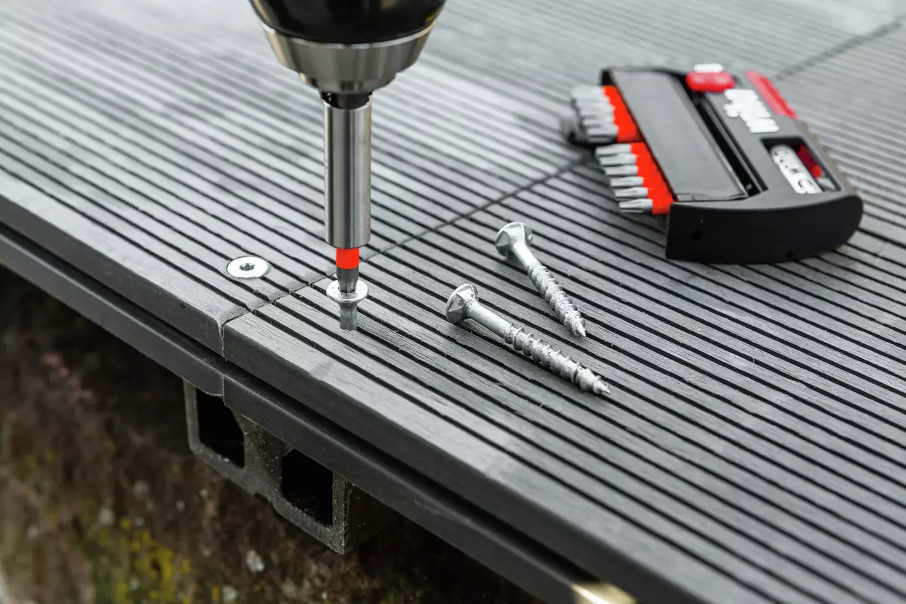 A screwdriver driving a screw into a grooved deck board, with extra screws and a bit set nearby.
