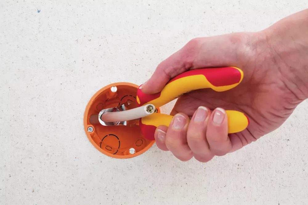 A person stripping a wire with red and yellow insulated pliers in an orange wall socket.