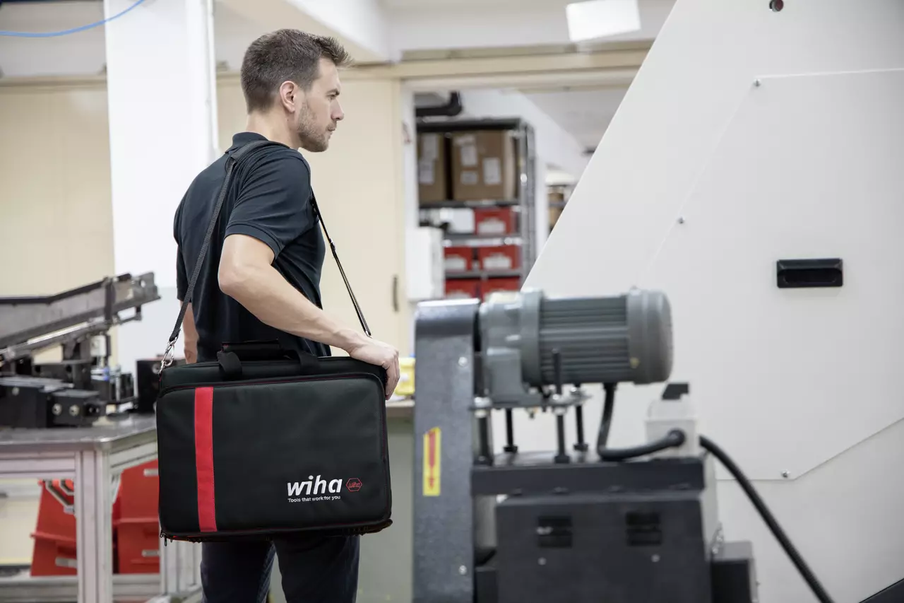 A man with a black bag in a workshop in front of a machine.