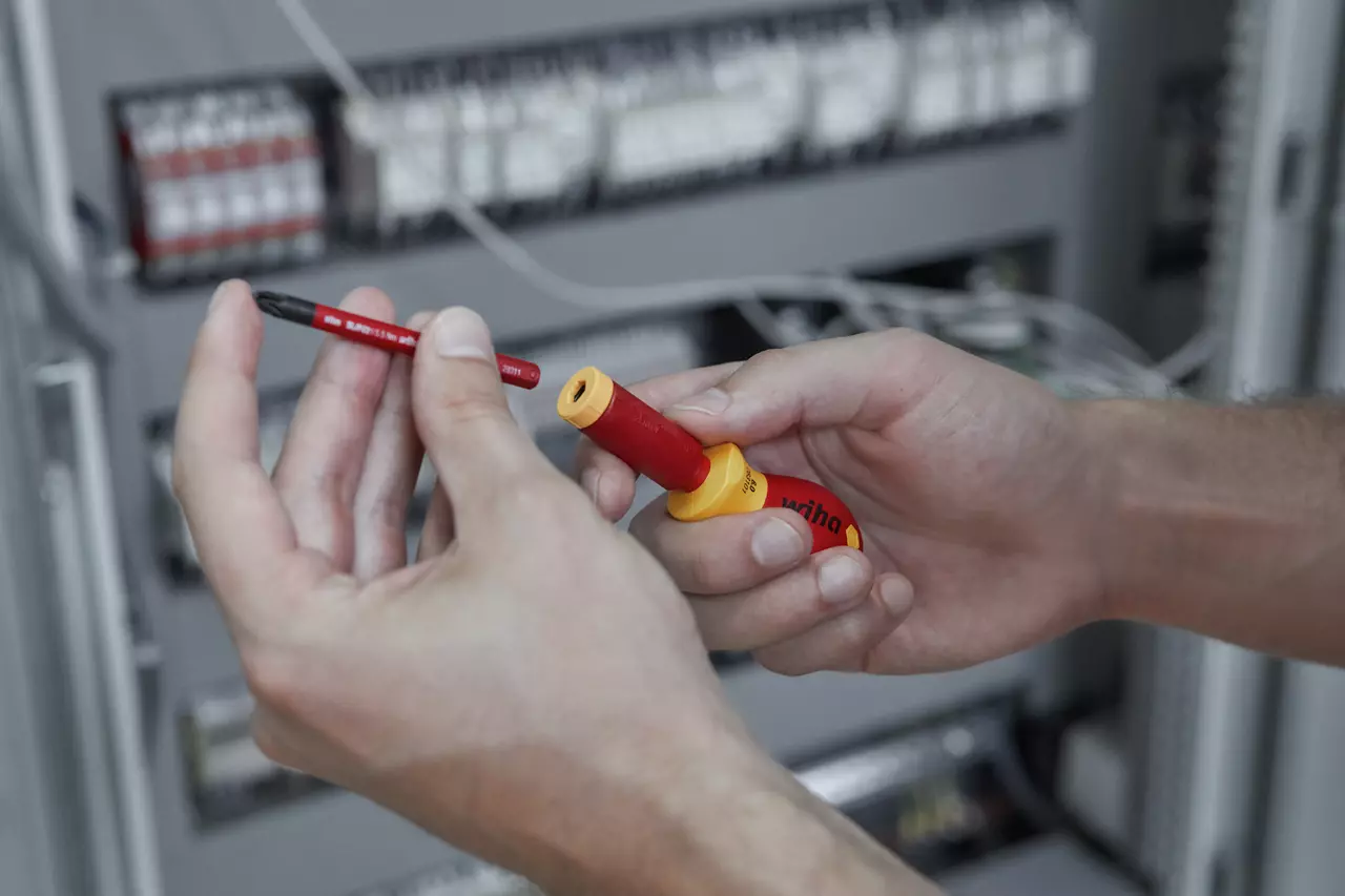 Person holding a red and yellow insulated screwdriver handle and blade in front of electrical panel.