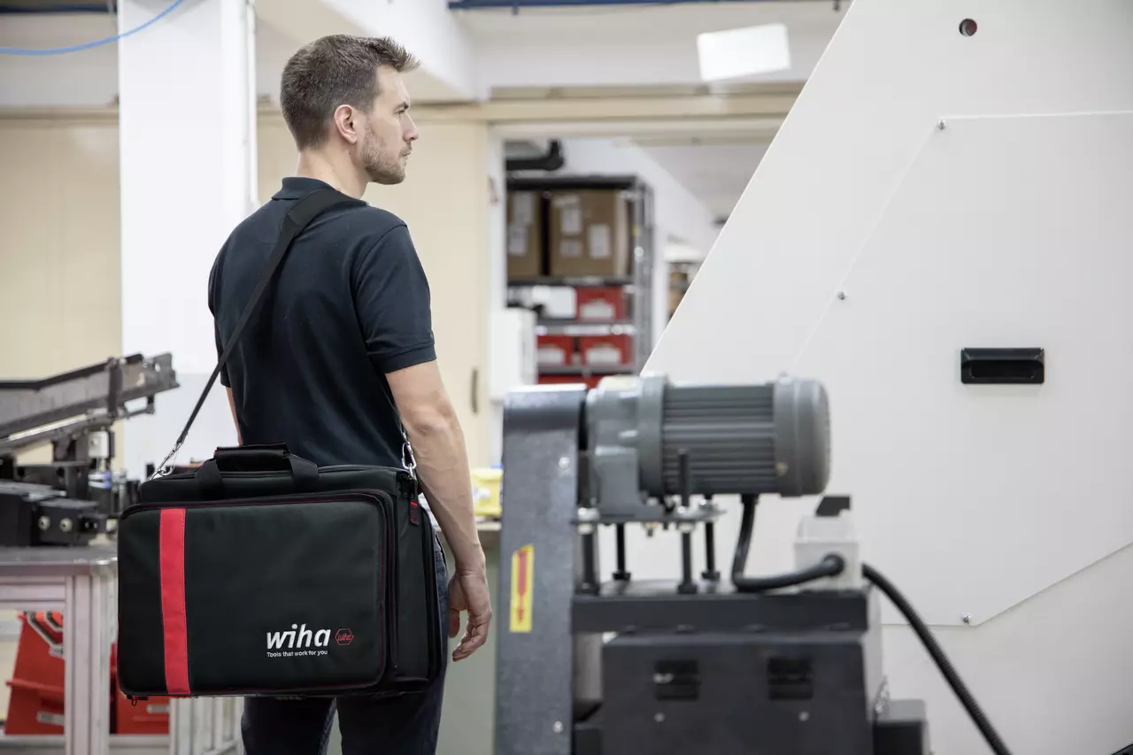 A person in a workshop carrying a black and red tool bag