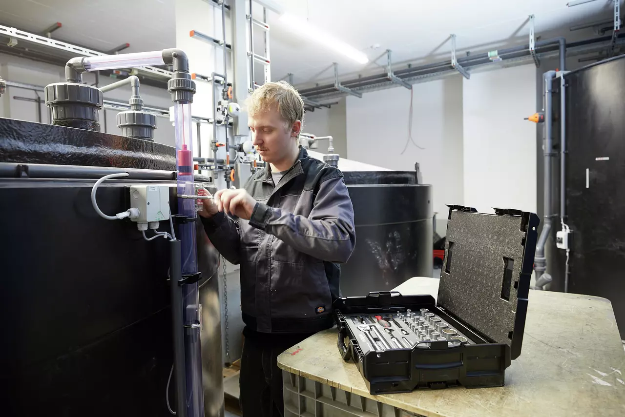 A person works on industrial pipes with tools from an open socket set case.