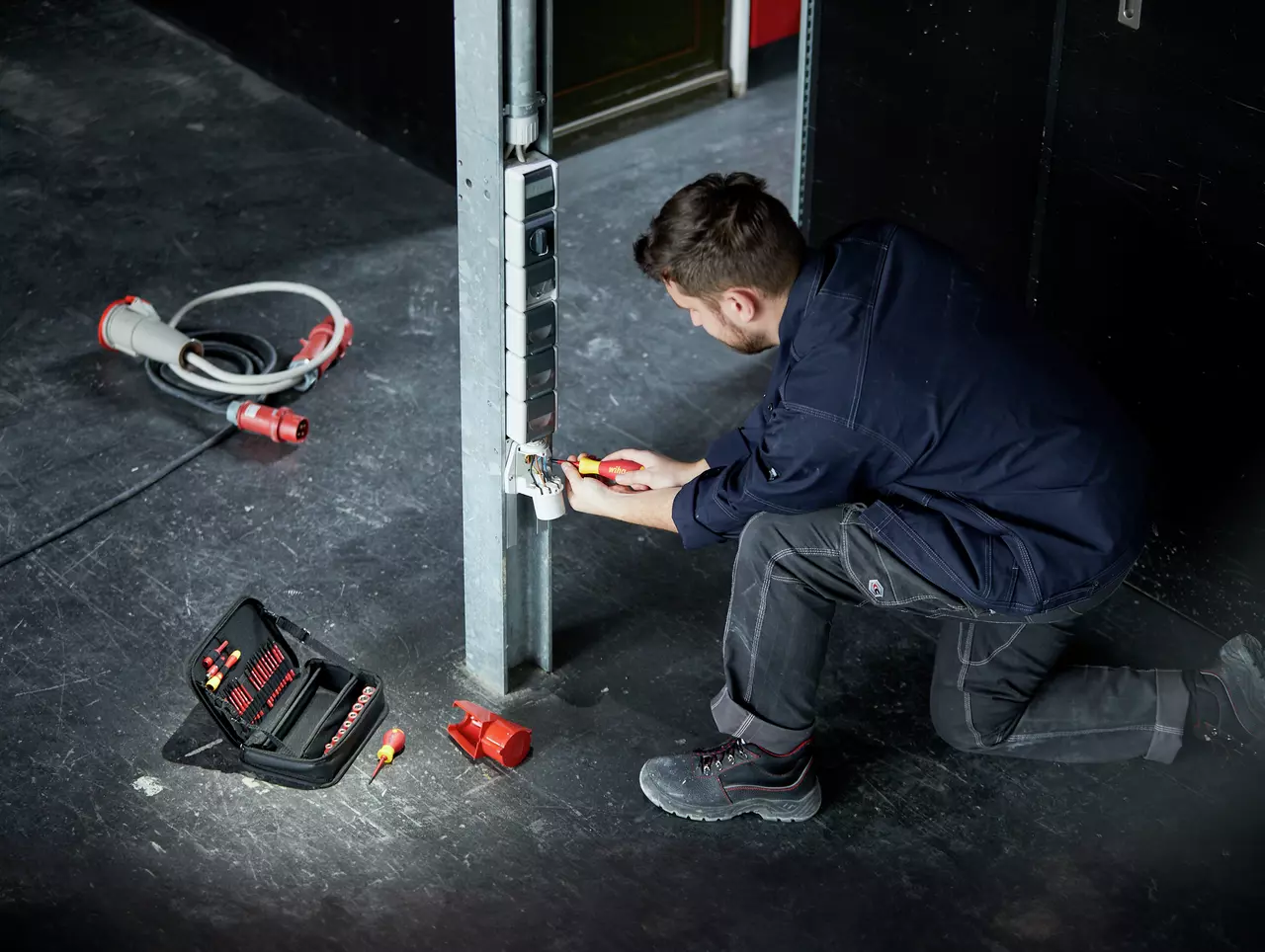 A person kneels, using a screwdriver on a power outlet. Tool kit and cables are on the floor.
