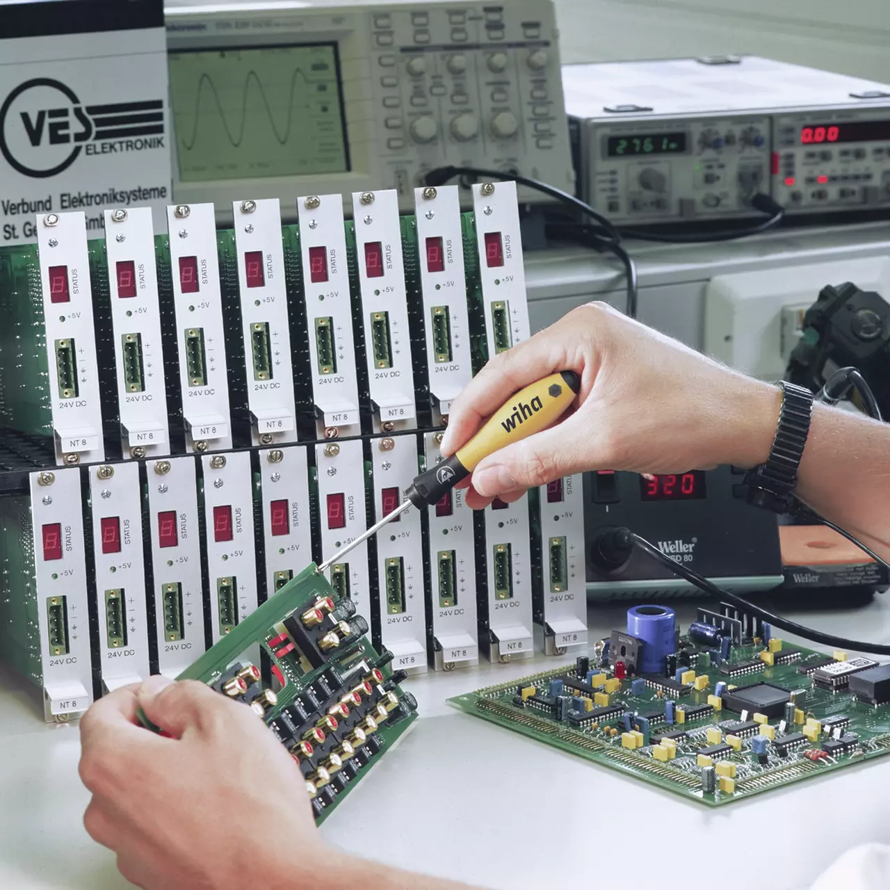 Person using an electrical screwdriver on a circuit board, surrounded by electronic testing equipment.