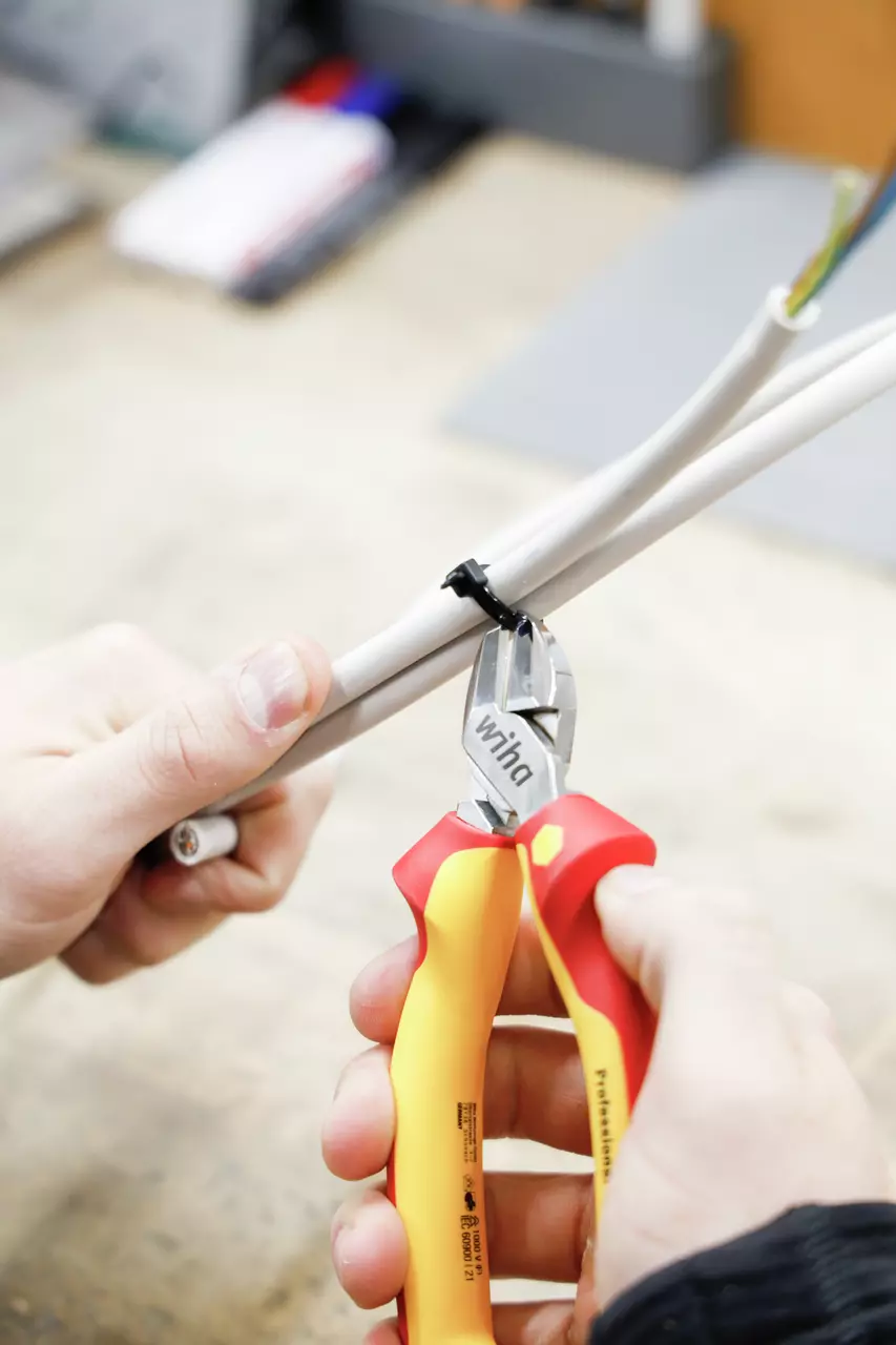 Person using red and yellow side cutter to cut a black cable tie on a white cable.