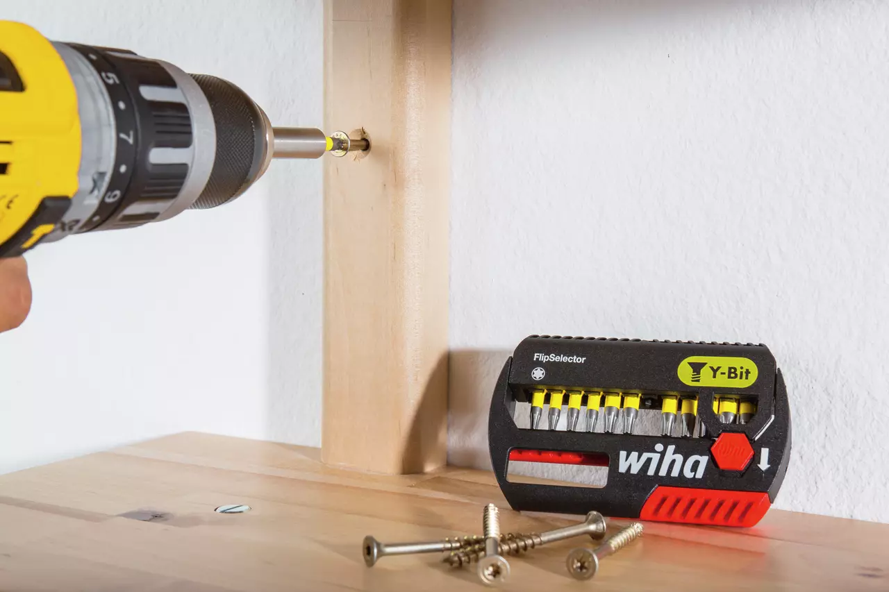 A person uses a yellow and black drill to drive a screw into a wooden surface, with a set of drill bits nearby.