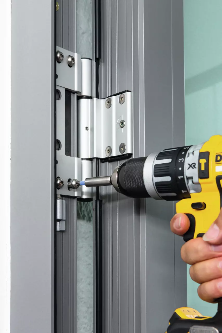 A person uses a yellow and black drill to adjust a metal hinge on a door frame.