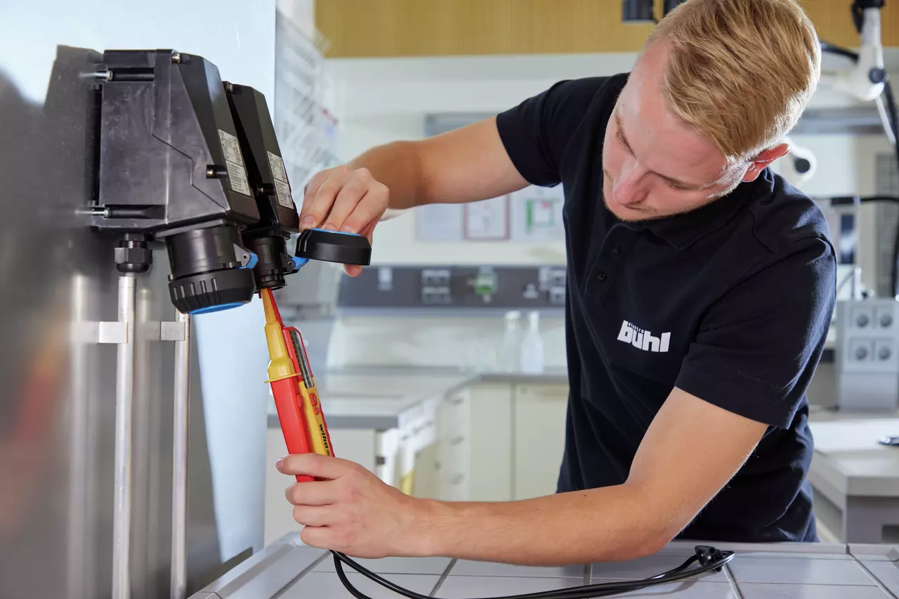 A person uses a voltage tester on a wall-mounted electrical device in a workshop.