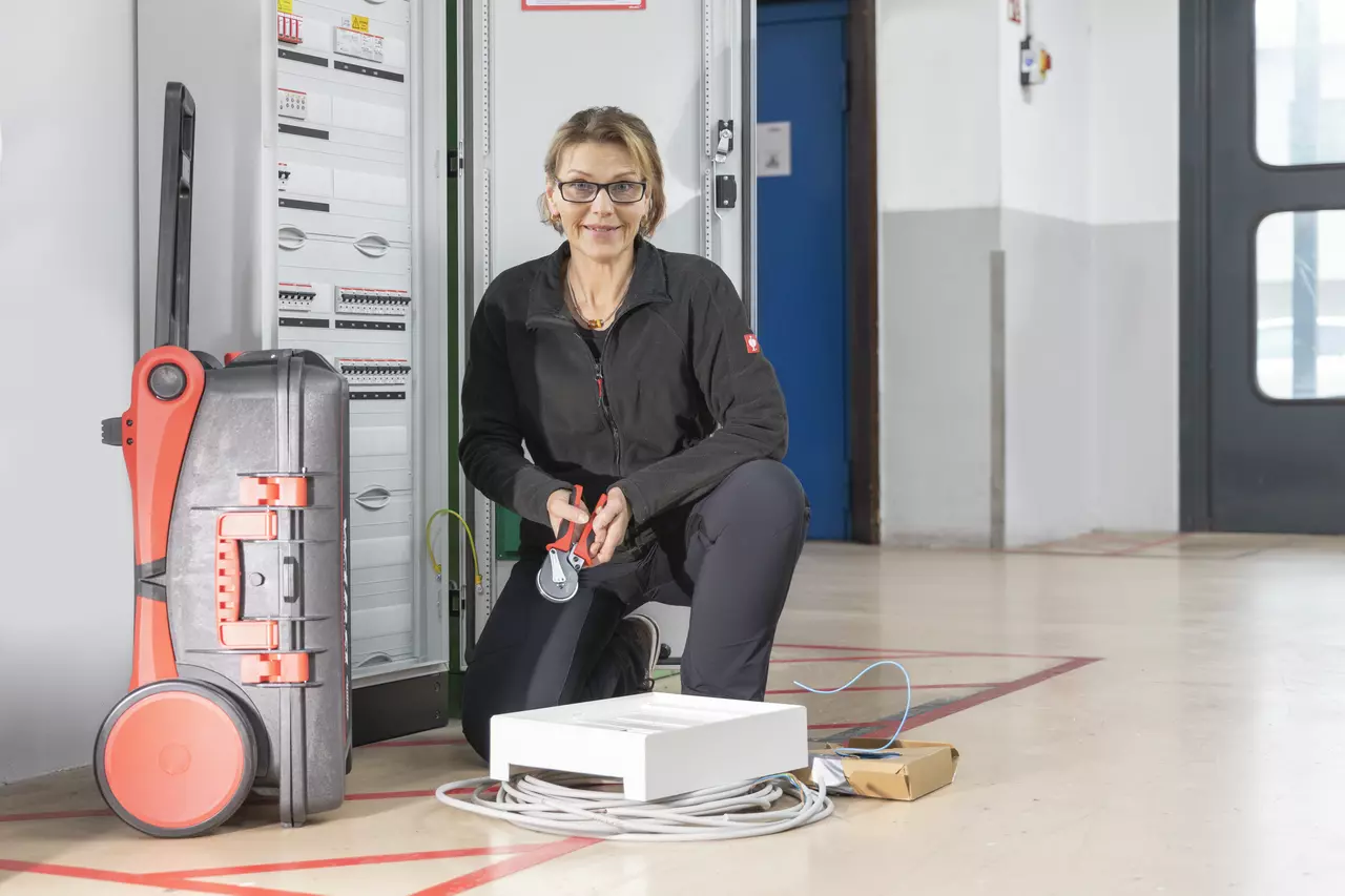 A person kneels in front of an electrical cabinet, holding a crimping tool.