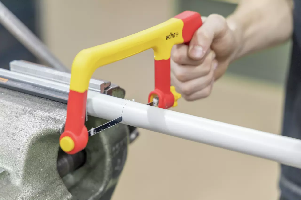 A person using a yellow and red hacksaw to cut a white pipe held in a vise.