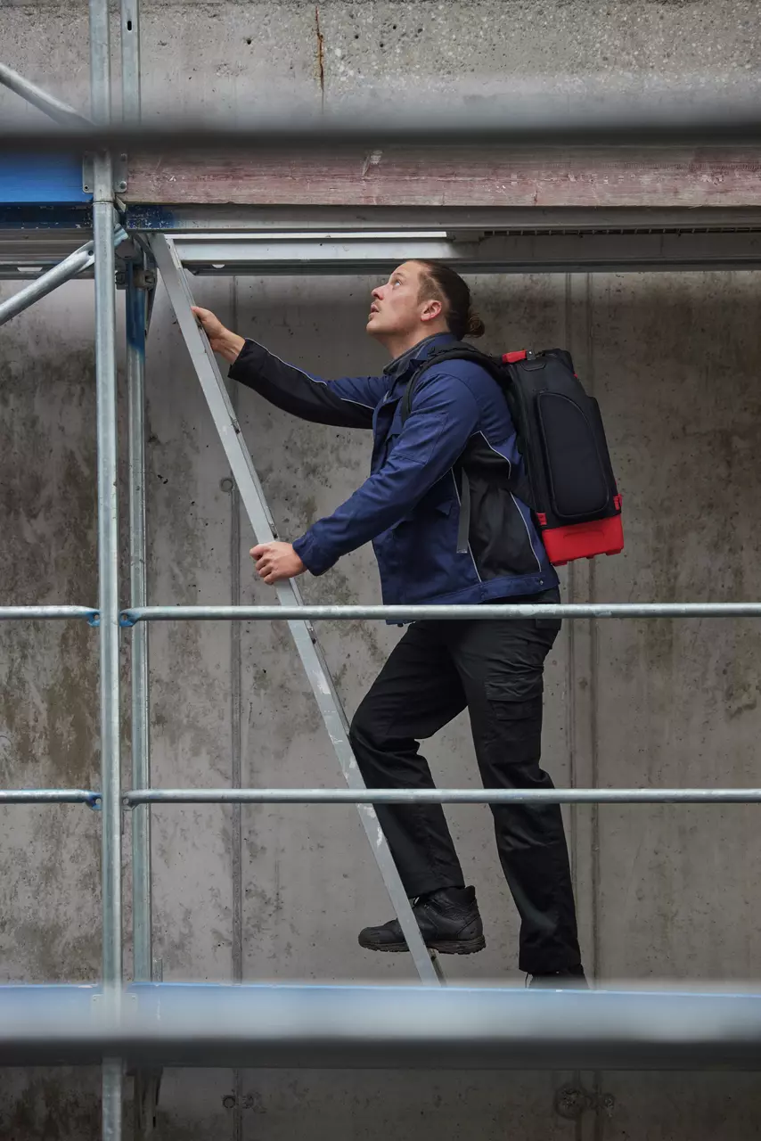 A person climbs a metal ladder on a scaffold, wearing a blue jacket and black pants.