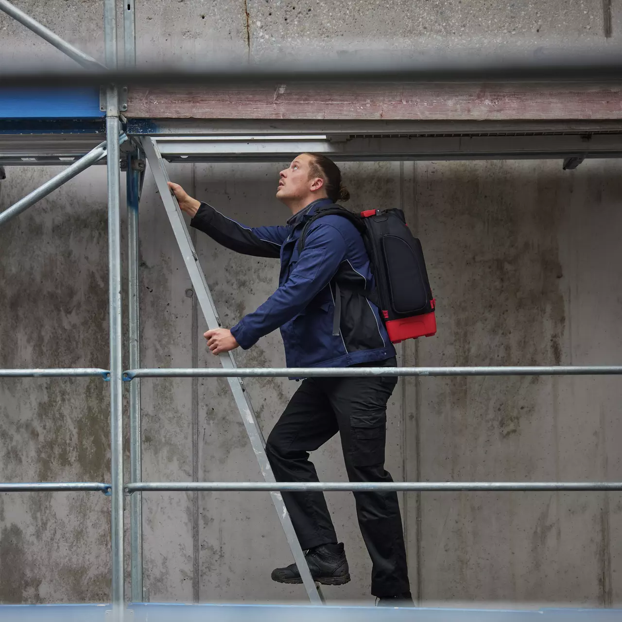 A person climbs a metal ladder on a scaffold, wearing a blue jacket and black pants.