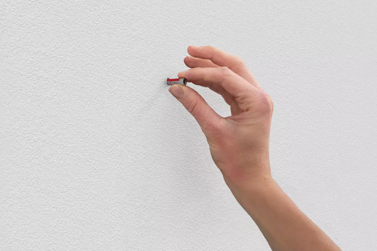 A hand holding a small tool against a white textured wall.