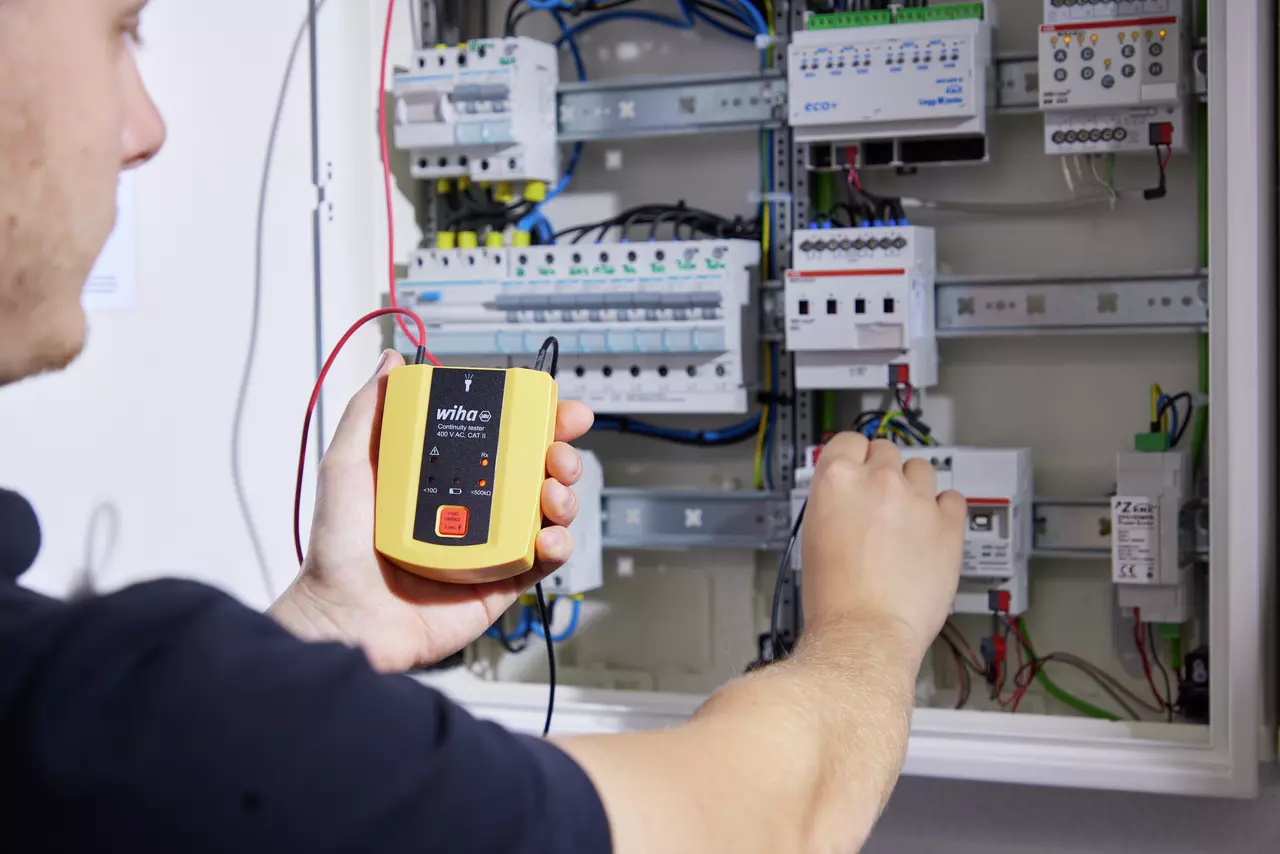 Person using a continuity tester on an open electrical control cabinet with wires and components visible.