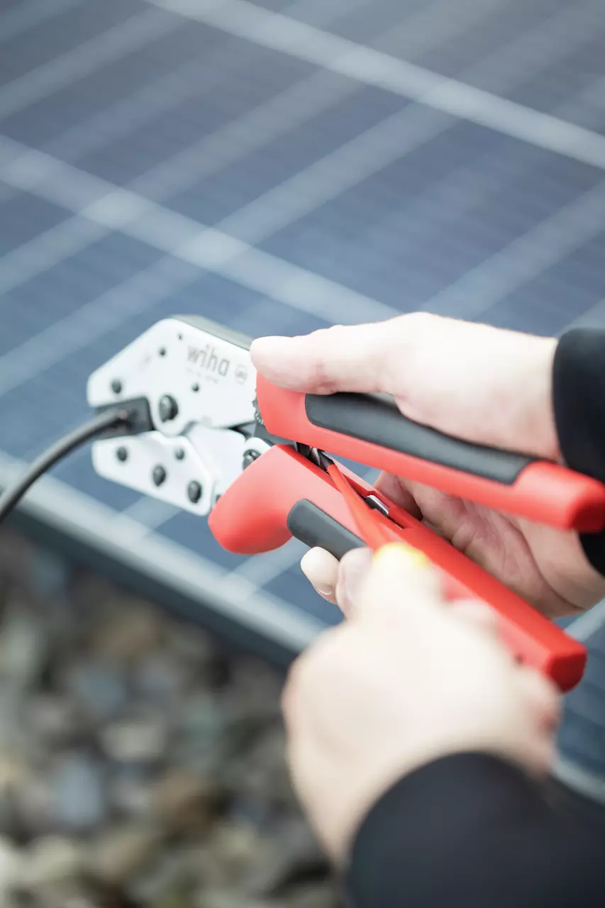 Person using a crimping tool with red handles on a cable near solar panels.