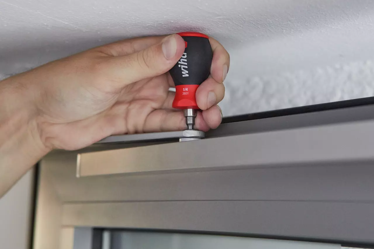 Hand using a red and black stubby screwdriver to adjust a screw on a metal door frame.