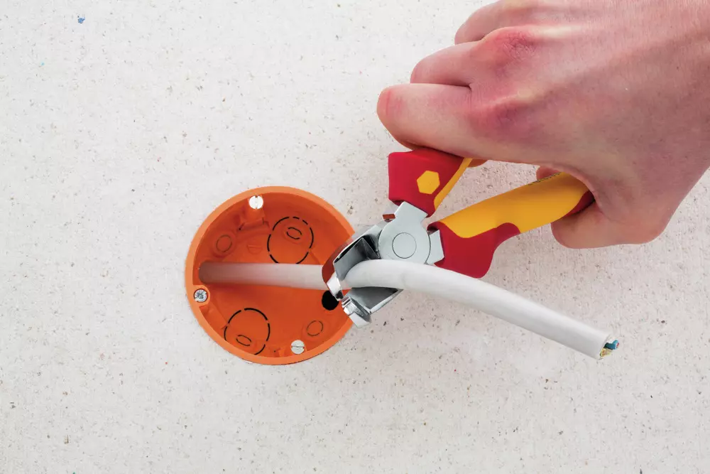 Hand using red and yellow wire cutter to cut white cable near orange outlet box.