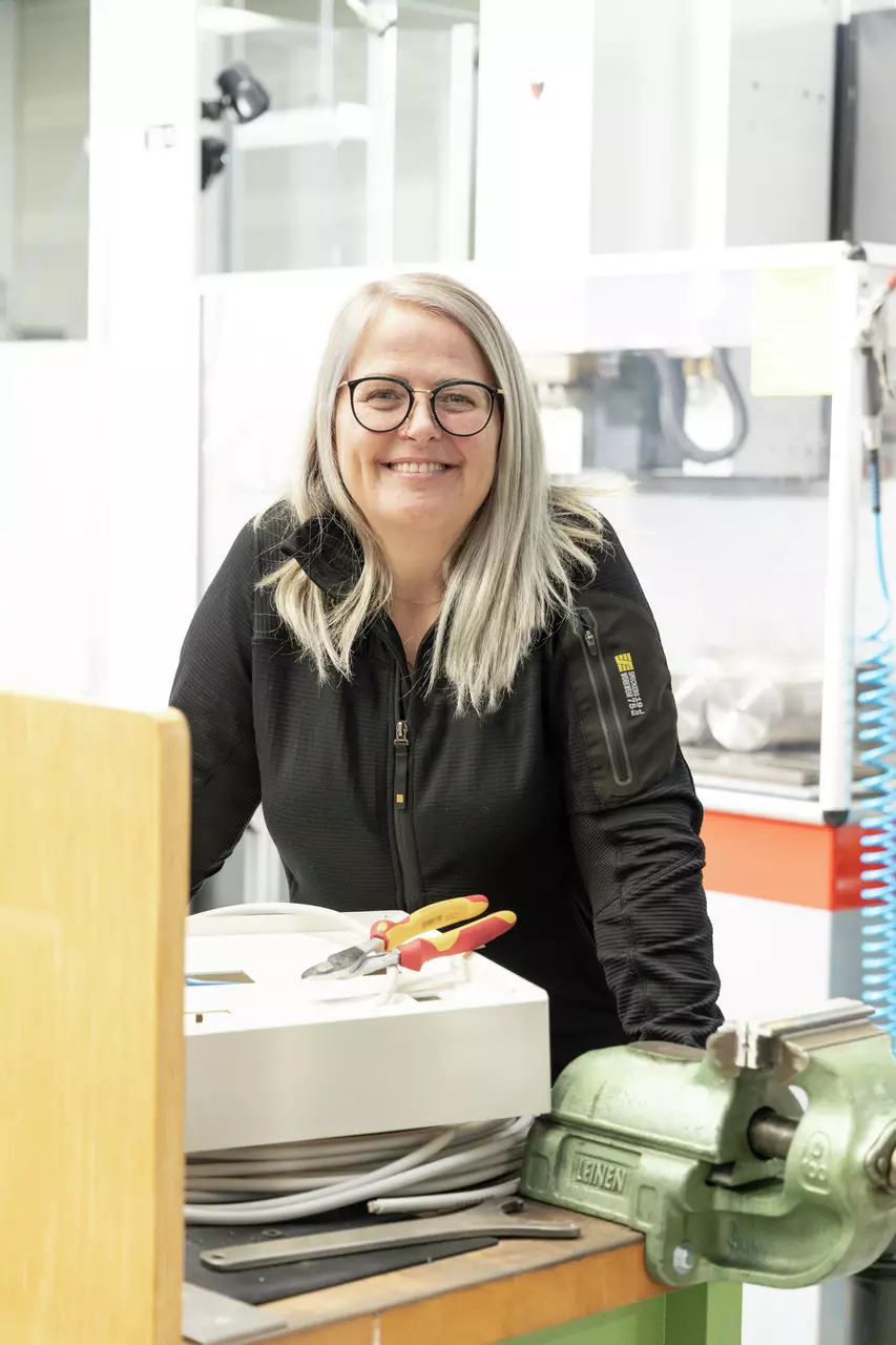 A person with glasses stands smiling in a workshop next to a vise and tools.