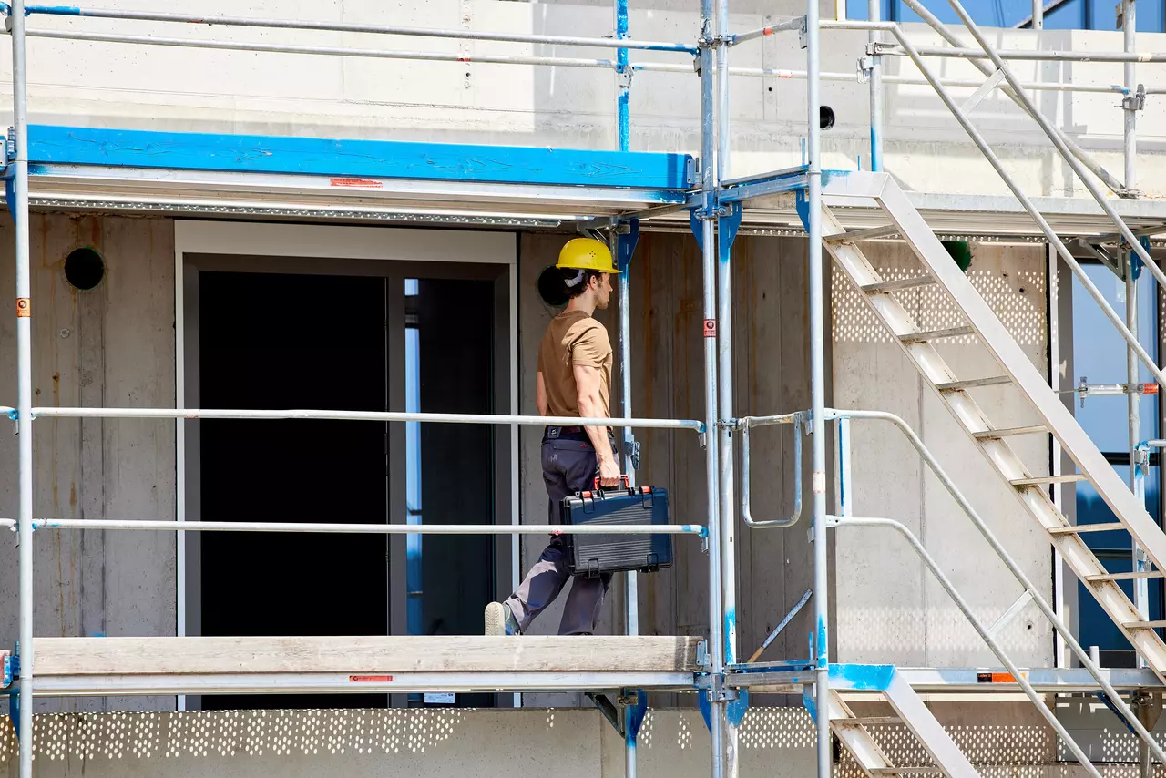 A person in a yellow hard hat carrying a toolbox on a construction site scaffold.