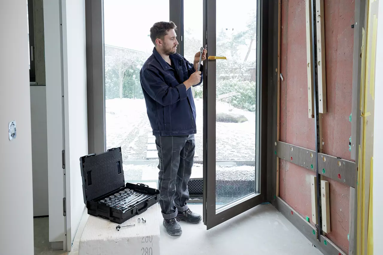 A person using a ratchet wrench on a window frame, with a socket set in a case nearby.