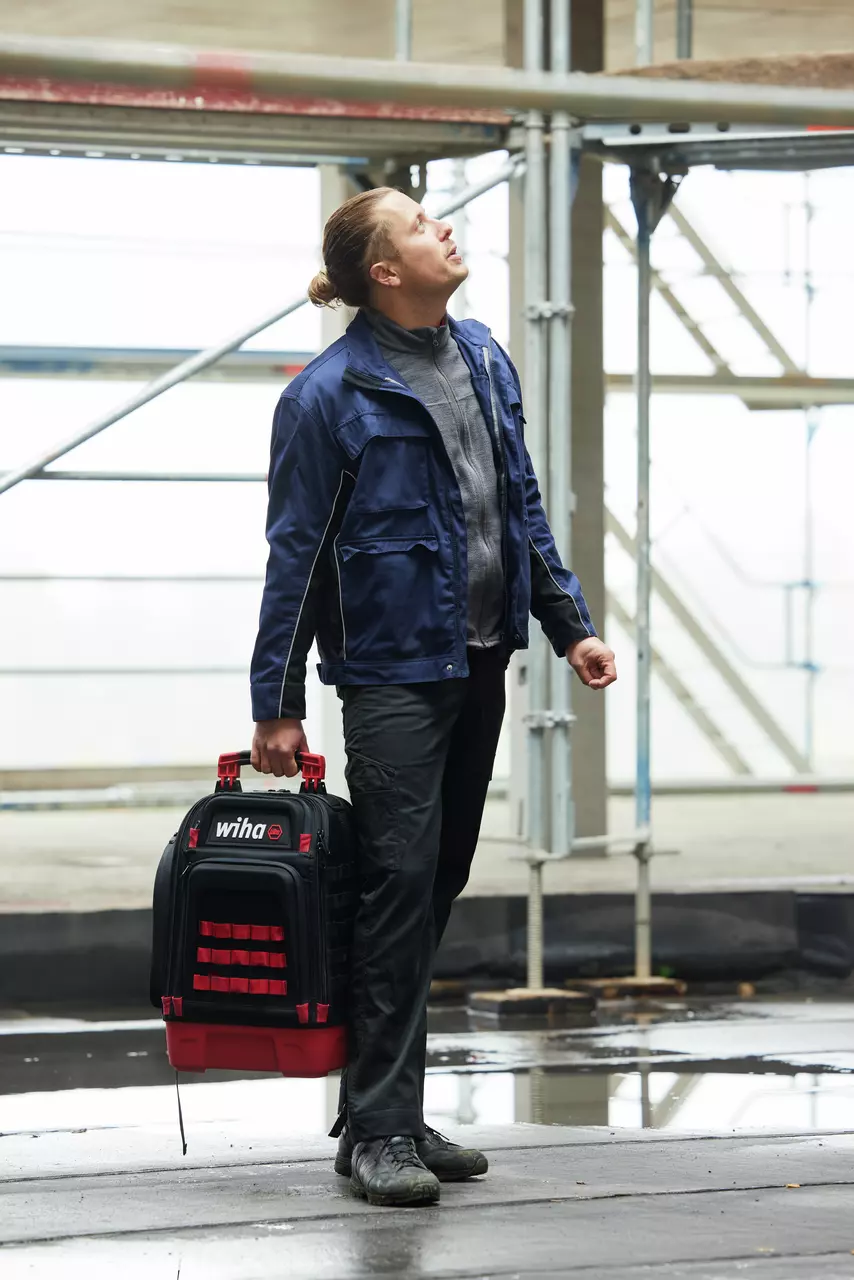 Person in workwear carrying a tool case and looking upwards, construction site environment.