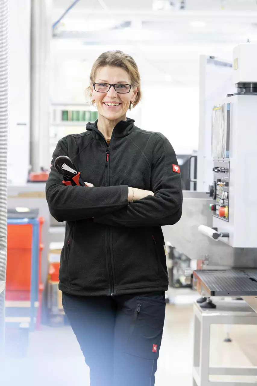 A smiling person in work attire stands in a workshop holding a tool.