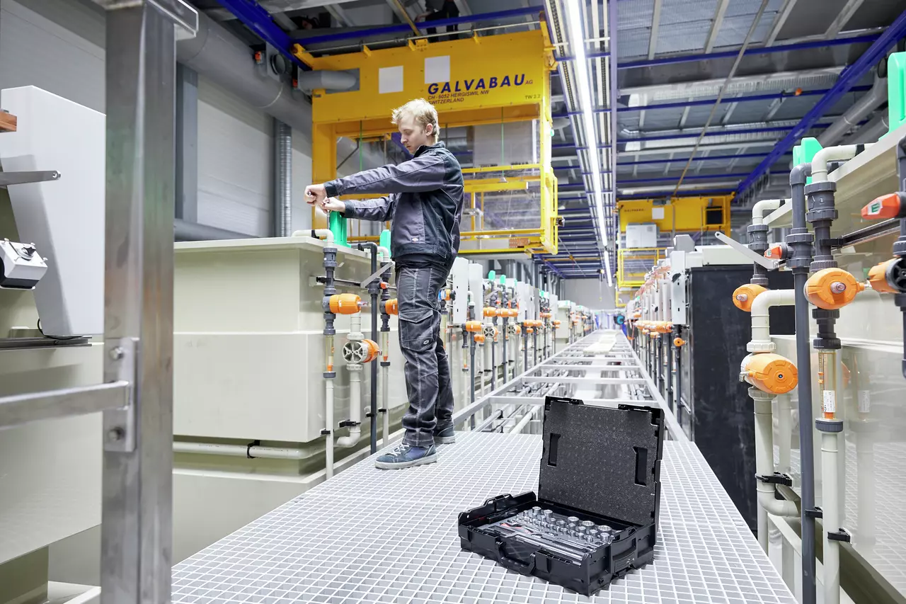 Open socket set case on industrial walkway, worker adjusting pipes in background.